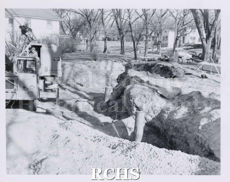 Black and white photograph of construction. Appears to be a trench with some poles sticking out of ground. Partial vehicle on left with driver.