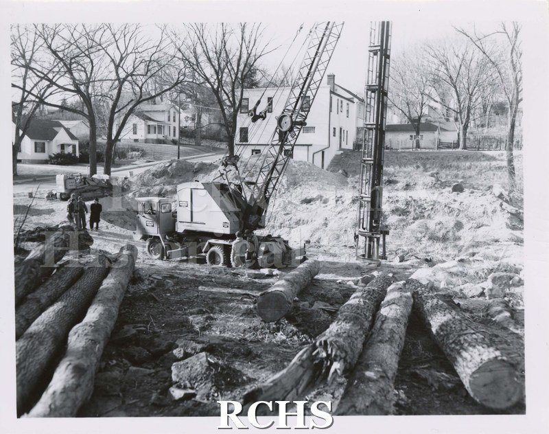 Black and white photograph of unidentified construction. Vehicle with shaft on right into hole. Two piles of logs with bark on each side of foreground.