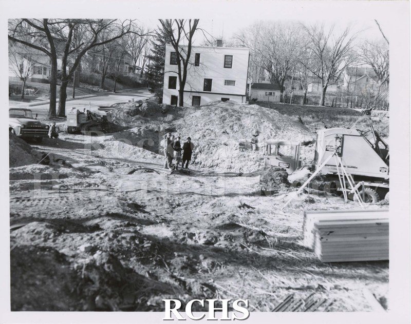 Black and white photograph of unidentified construction. Partial vehicle with shaft on right. Stack of lumber on front right. Four men standing at center.