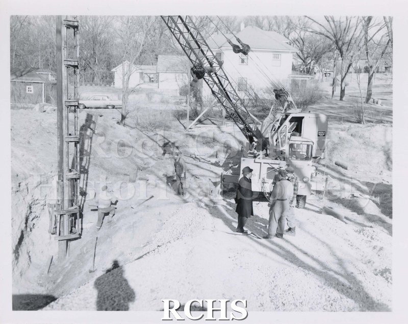 Black and white photograph of unidentified construction. Vehicle with shaft in hole. One man climbing from hole, another man looking down at him.