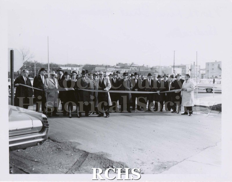 Black and white photograph of North River Street Parking Lot Dedication. Shows several people standing behind a ribbon on a parking lot.