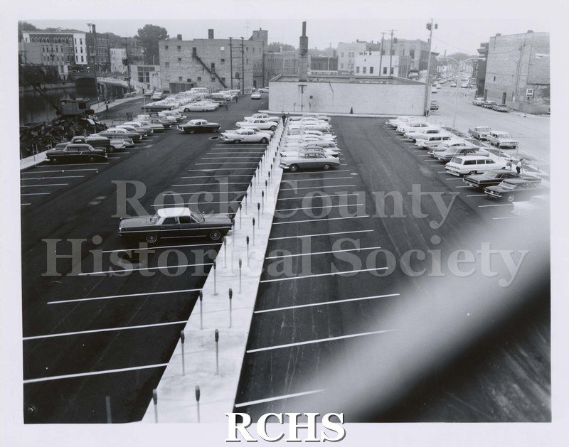 Black and white photograph of North River Street Parking Lot with cars parked on it. Buildings are visible in the background.
