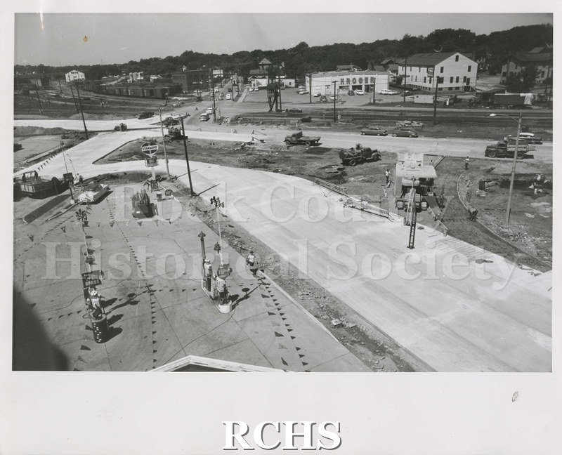 Black and white photograph of construction site. The road is paved. On the left side is the Standard Gas Station; there is construction equipment on the right.