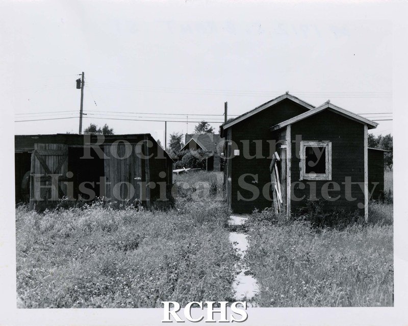 Black and white photograph of 1712 S. Grant St., Janesville, WI. Shows a small house with a shed beside it. Grass has grown up around the walkway.