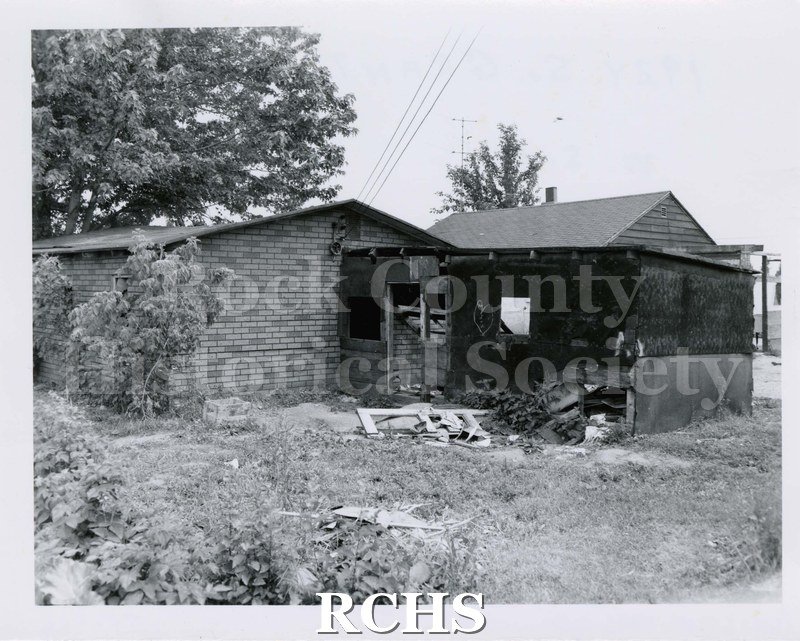 Black and white photograph of 1924 S. Grant St., Janesville, WI. Shows an abandoned brick building with a structure in front. There is debris around the house.