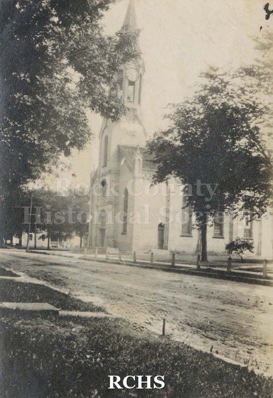 Black and white photograph of St. Patrick's Church. Shows a fence in front of the church with trees on the side.