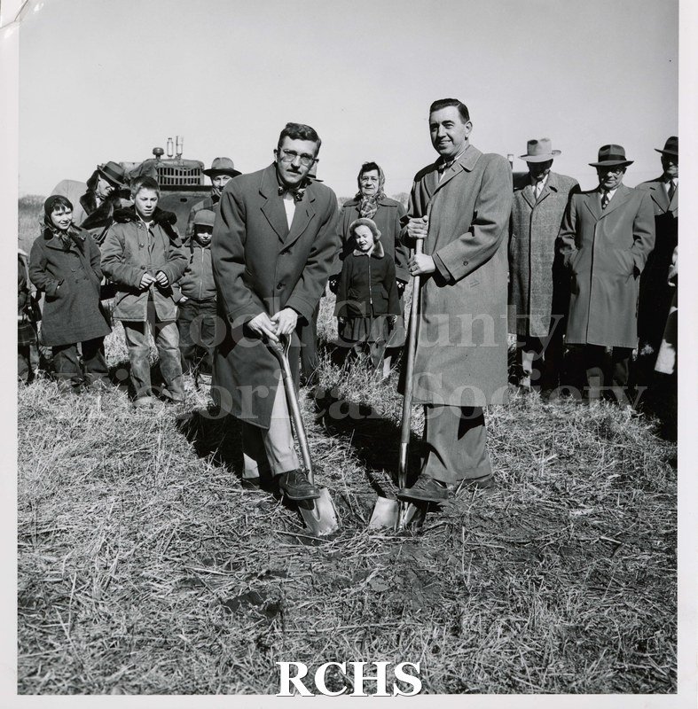 TPhotograph. Black and white photograph of two men, each with a foot on a shovel ready to break ground. There are several people standing behind them.