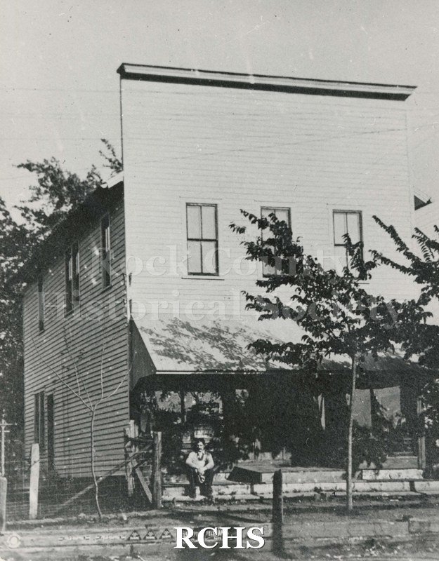 Black and white photograph of Springbrook Grocery store with an awning and false front. There is a man sitting on the steps.