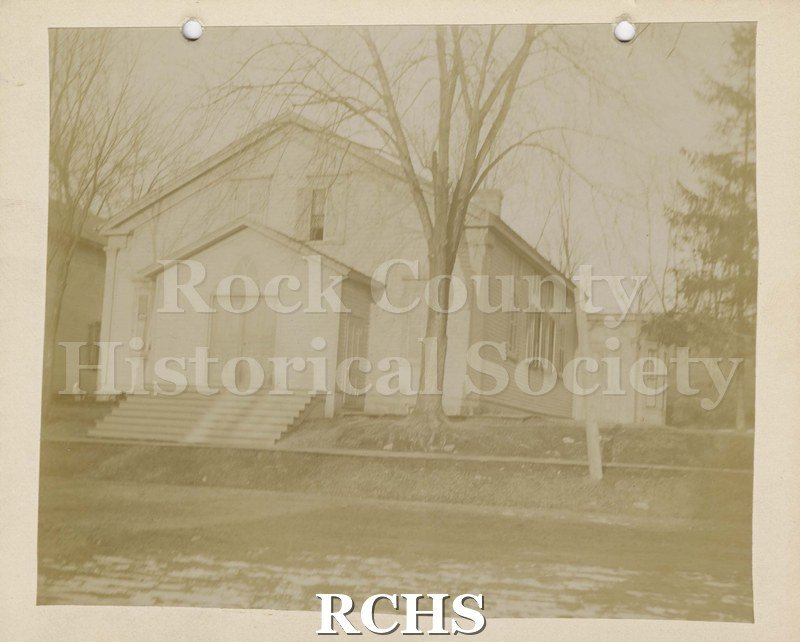 Black and white photograph of German Lutheran, (St. John's), Janesville, WI, an old wooden church building with wooden steps going up to the wooden doors.