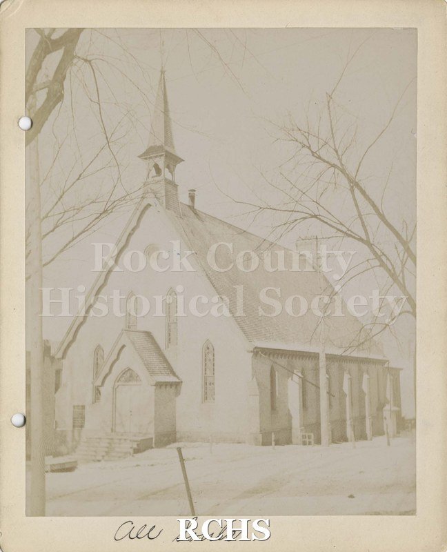 Black and white photograph of All Soul's Church, Janesville, WI, a building with a steeple on top and with four stained glass windows in the front.