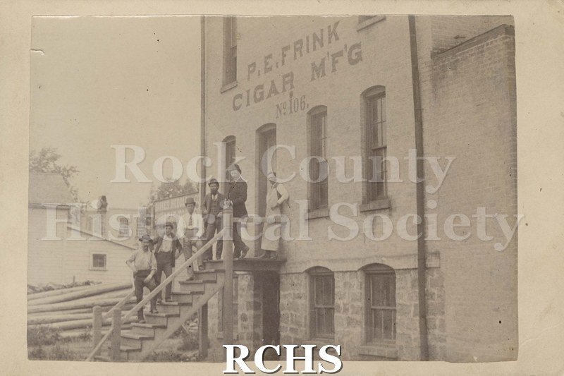 Black and white photograph of six unidentified men standing on the steps of a cigar warehouse. The sign on the building reads: "P. E. Frink Cigar M'F'G No. 106".