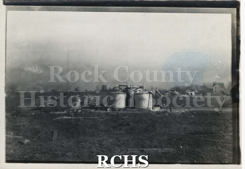 Black and white photograph of the tornado 1911. Shows oil tanks damaged by a tornado. There is a house standing on the right.