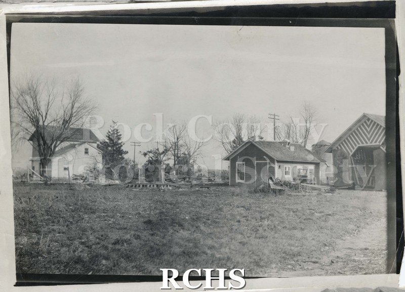 Black and white photograph of tornado damage to a house. Outbuildings are still standing after a tornado. There is debris from downed trees.