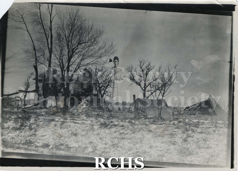 Black and white photograph of a tornado in Rock County 1911. Shows a farm with house intact but outbuilding on right demolished. Windmill is standing beside house.