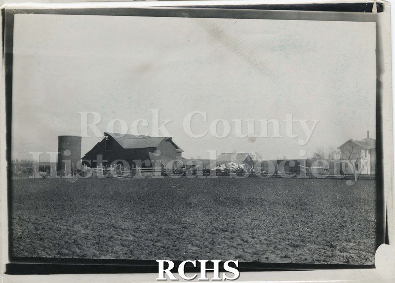 Black and white photograph of a tornado in Rock County. Shows a farm with the barn and other structure demolished. House on right appears intact.