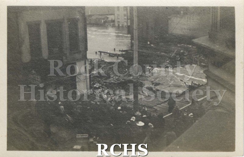 Black and white photograph of Milwaukee Street Fire 1913. Shows debris from the fire with a building on the left with a door and two windows.