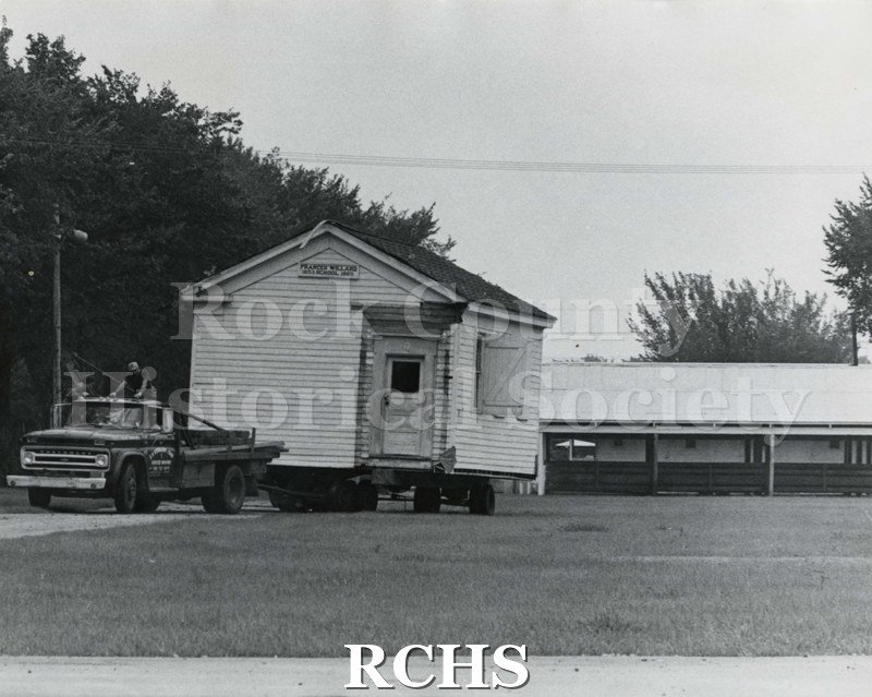 Black and white photograph of the Frances Willard Schoolhouse moving to fairgrounds. Truck has a driver and a man standing behind the cab.