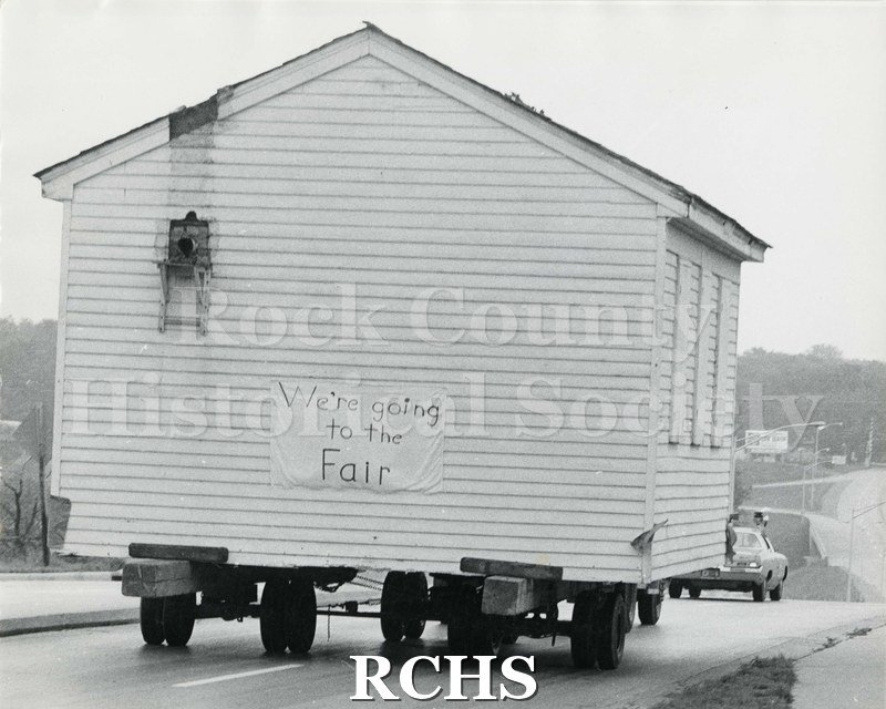 Black and white photograph of the Frances Willard Schoolhouse moving to fairgrounds. It is being moved along the road.