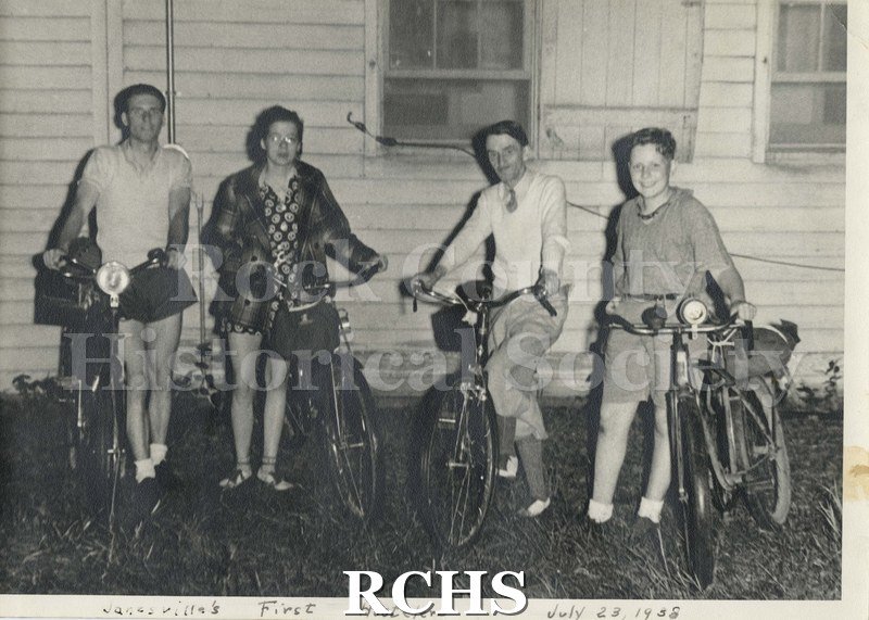 Black and white photograph of Frances Willard School hostel visitors. Shows three men and one woman standing beside their bikes in front of a building.