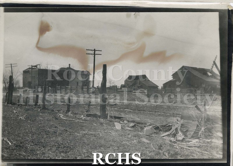 Black and white photograph of damage from tornado of 1911.Barn on right side has roof torn off and a light pole is toppled.