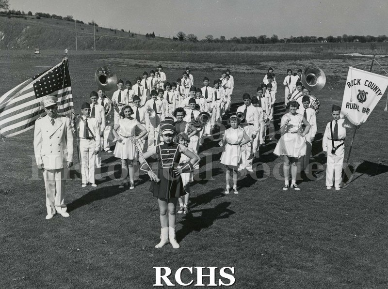 Black and white photograph of Rock County Rural Band 1954 lined up on a field. The boy on the left is holding a banner for: "Rock County Rural Band".