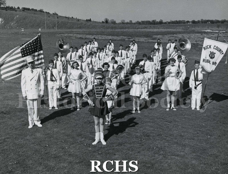 Black and white photograph of Rock County Rural Band lined up on a field. The boy on the left is holding a banner for: "Rock County Rural Band".