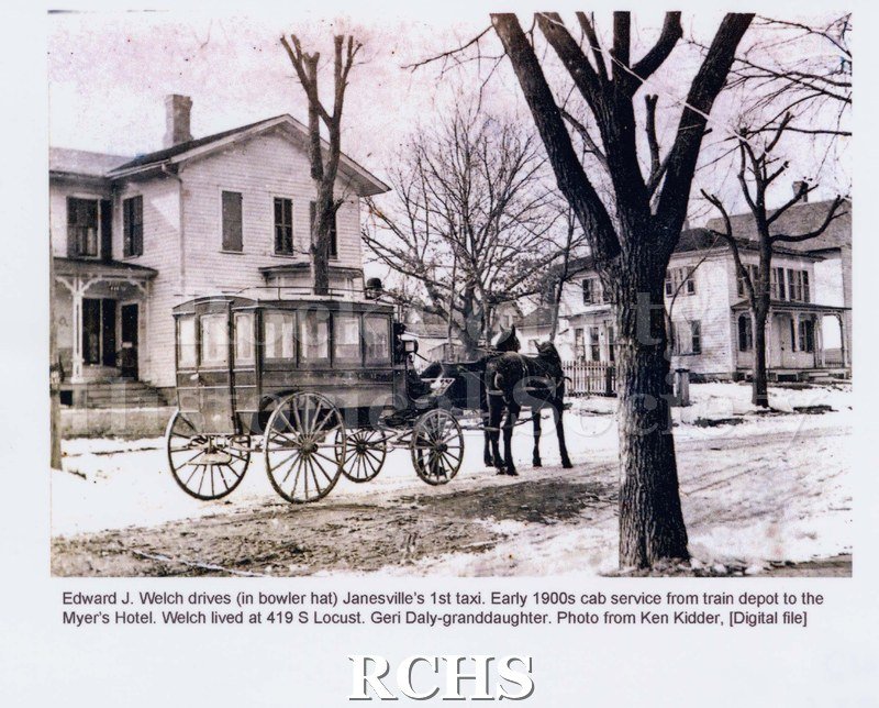 Black and white photograph of Edward J. Welch driving Janesville's 1st taxi, early 1900s. Shows a carriage pulled by two horses in snow covered street.