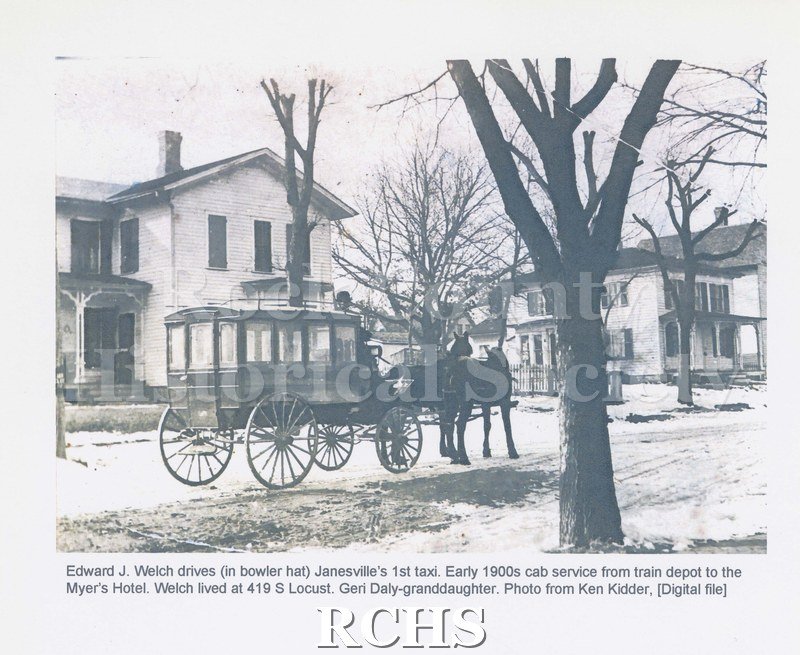 Black and white photograph of Edward J. Welch driving Janesville's 1st taxi, early 1900s. Shows a carriage pulled by two horses in snow covered street.