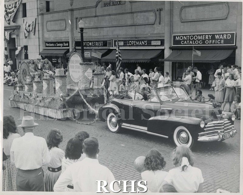Black and white photograph of Swanson Grocery Parade Float. Shows car pulling parade float.