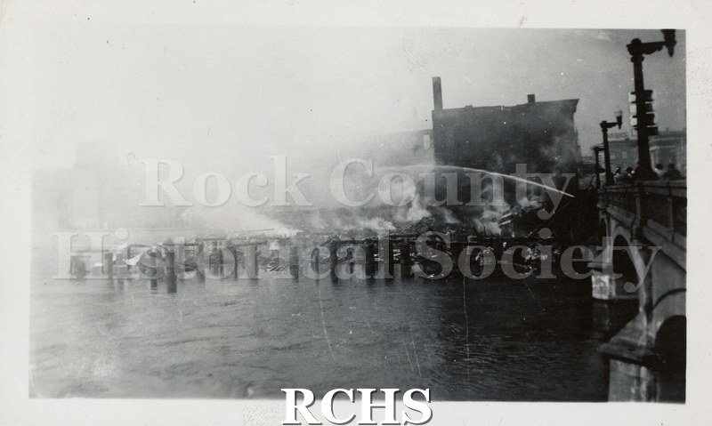 Black and white photograph of Milwaukee Street Bridge Fire, 1941.. From right side of photograph, a stream of water coming from concrete/stone bridge with light.