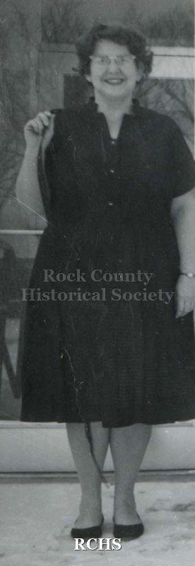 Black and white photograph of an unidentified woman standing outside the entrance to a building, wearing a dark-colored dress. Undated.