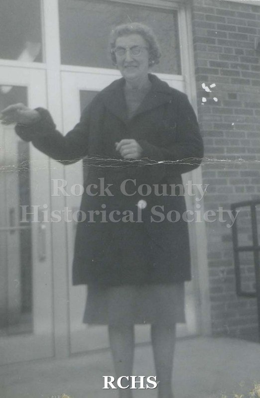 Black and white photograph of an unidentified woman standing outside the entrance to a building, wearing a skirt and dark-colored jacket. Undated.