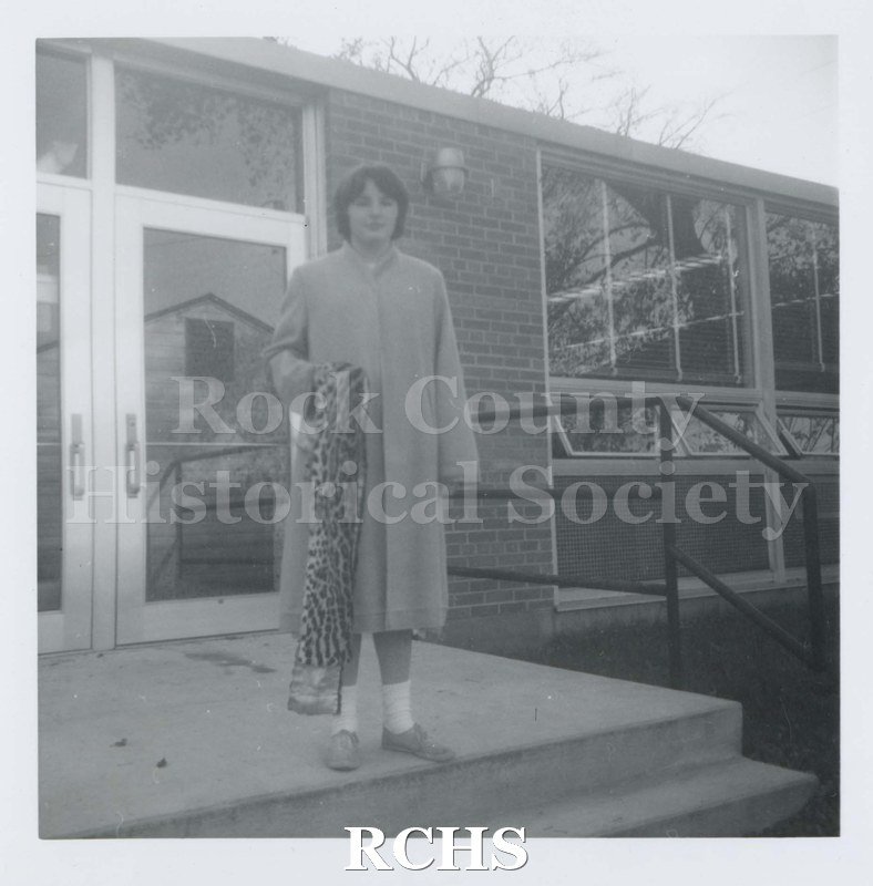 Black and white photograph of an unidentified woman standing outside the entrance to a building, wearing a long coat and holding a piece of leopard-printed fabric.
