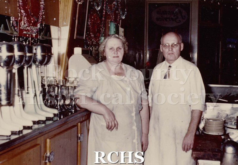Color photo of Howard Took and wife behind the counter of the ice cream store. There are malted milk makers on the counter beside them.
