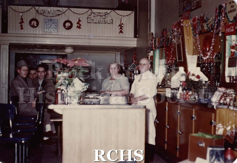 Color photo of Howard Took and his wife behind the counter in his store. There are customers on the left and a large poinsetta plant on the table in the front.