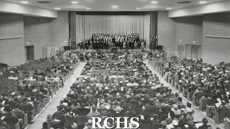 Black and white photograph of choir in robes on stage with many adults sitting in front of them. The orchestra is in the pit in front of the stage.