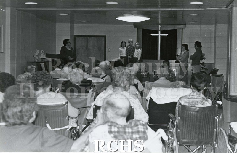 Black and white photograph of the interior of Rock County Health Care Center, a chapel with a cross, with three children performing on the stage.