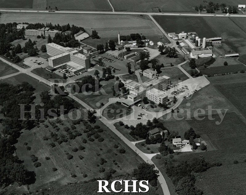 Black and white photograph of an aerial photograph of the Rock County Health Care facility. Shows dairy barns on top right and other buildings.