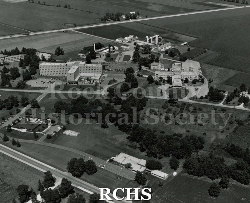 Black and white photograph of an aerial photograph of the Rock County Health Care facility. Shows dairy barns on top right and other buildings.