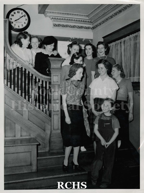 Black and white photograph of ten unidentified adults and children standing on a stairway. There is a clock on the wall behind them.