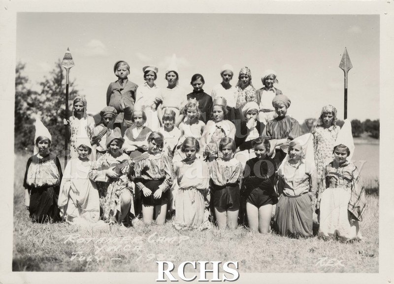 Black and white photograph of several people sitting and standing in a field. All are wearing costumes.
