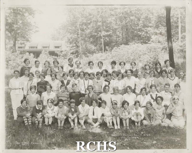 Black and white photograph of several people sitting and standing in a forested area. On bottom: "Beloit-Janesville YWCA Lake Geneva 1928".
