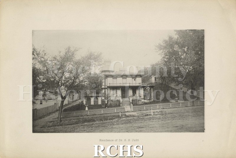 Black and white photograph of the residence of Dr. S. S. Judd. Shows a two story house with a picket fence in front of it. On bottom: "Residence of Dr. S. S. Judd".