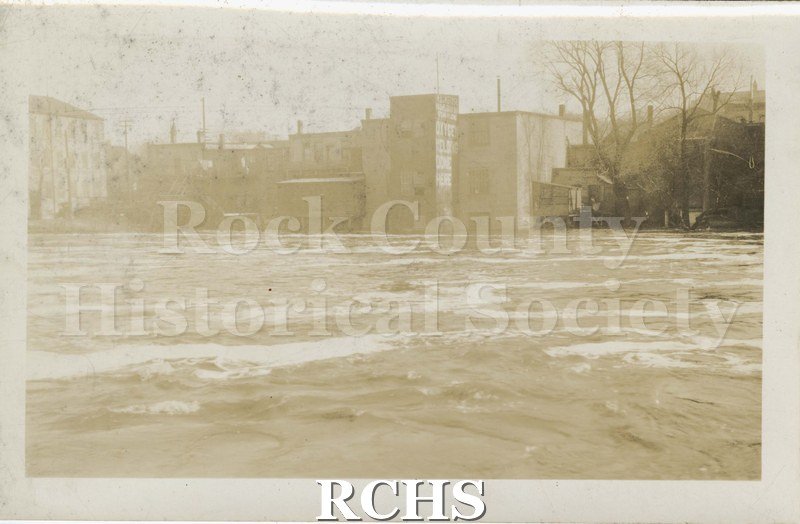 Black and white photograph of Rock River at flood stage. Several buildings are in the background.