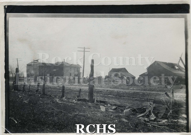 Black and white photograph of the Rock County tornado 1911. Shows a farm that had tornado damage. The barn on the right has the roof torn off.