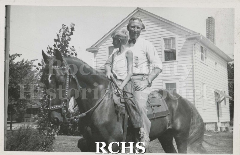 Black and white photograph of Chester Welch sitting on a horse with a boy in front of him.They are posing in front of a house.