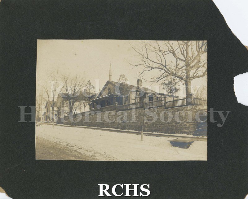 Black and white photograph of 218, 228, 302 Prospect Avenue, Janesville, WI. Shows a house with a fence in front of it which is built on a stone wall.