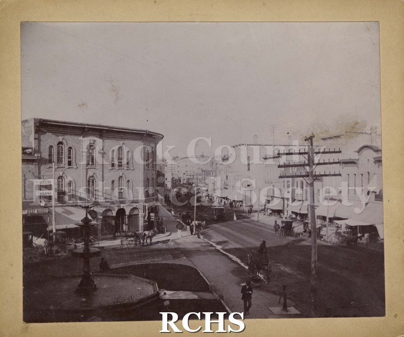 Black and white photograph of Milwaukee Street, Janesville, WI., showing an intersection and horses and buggies on the street.