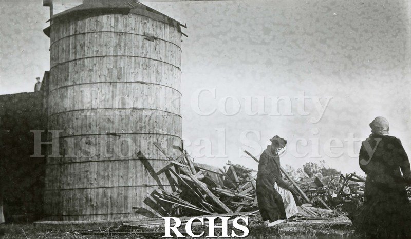 Black and white photograph of damage from tornado of 1911. Silo is standing but rubble in in front. Two women are pictured looking at the rubble.