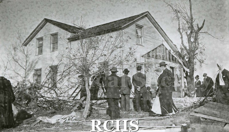 Black and white photograph of damage from tornado of 1911. Several people are standing looking at the damage to a farmhouse.